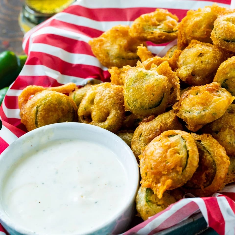Crispy fried pickles in a red and white striped basket with a side of ranch dipping sauce.