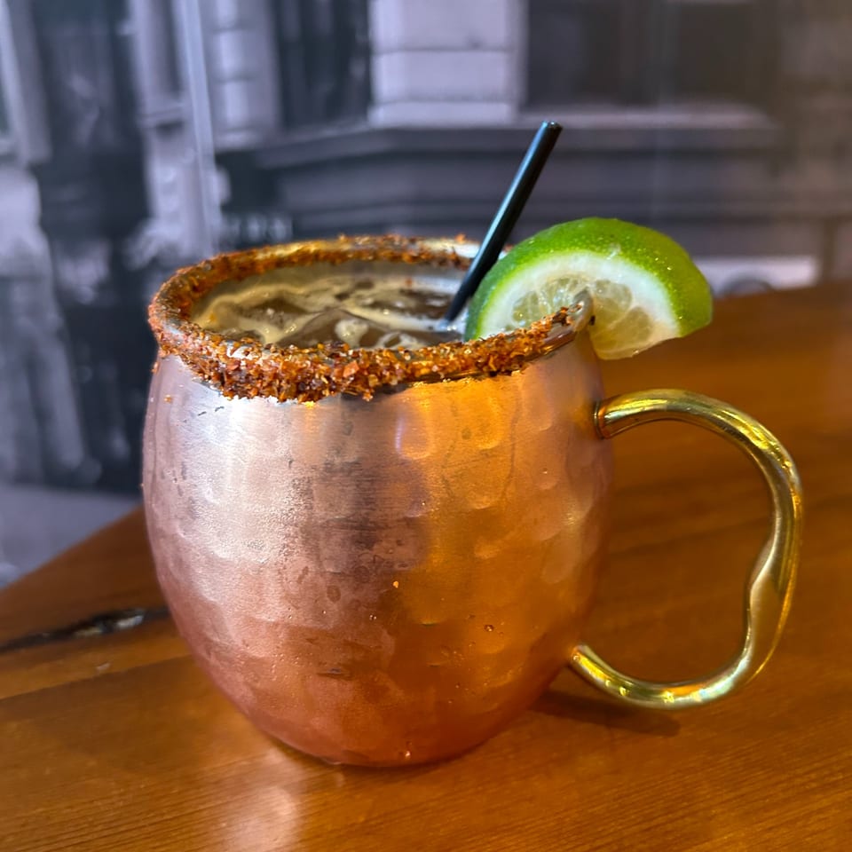 A copper mug with a spiced rim, dark drink, black straw, and lime wedge on a wooden table.