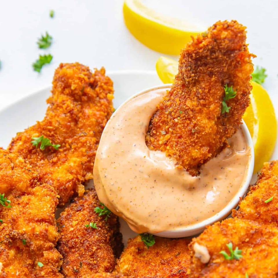 Crispy breaded chicken tenders with a creamy dipping sauce, garnished with parsley and lemon wedges.