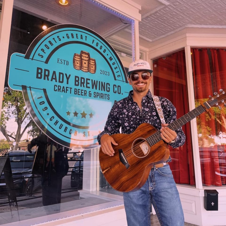 A man with a guitar stands outside Brady Brewing Co., a brewery with a blue and white sign.