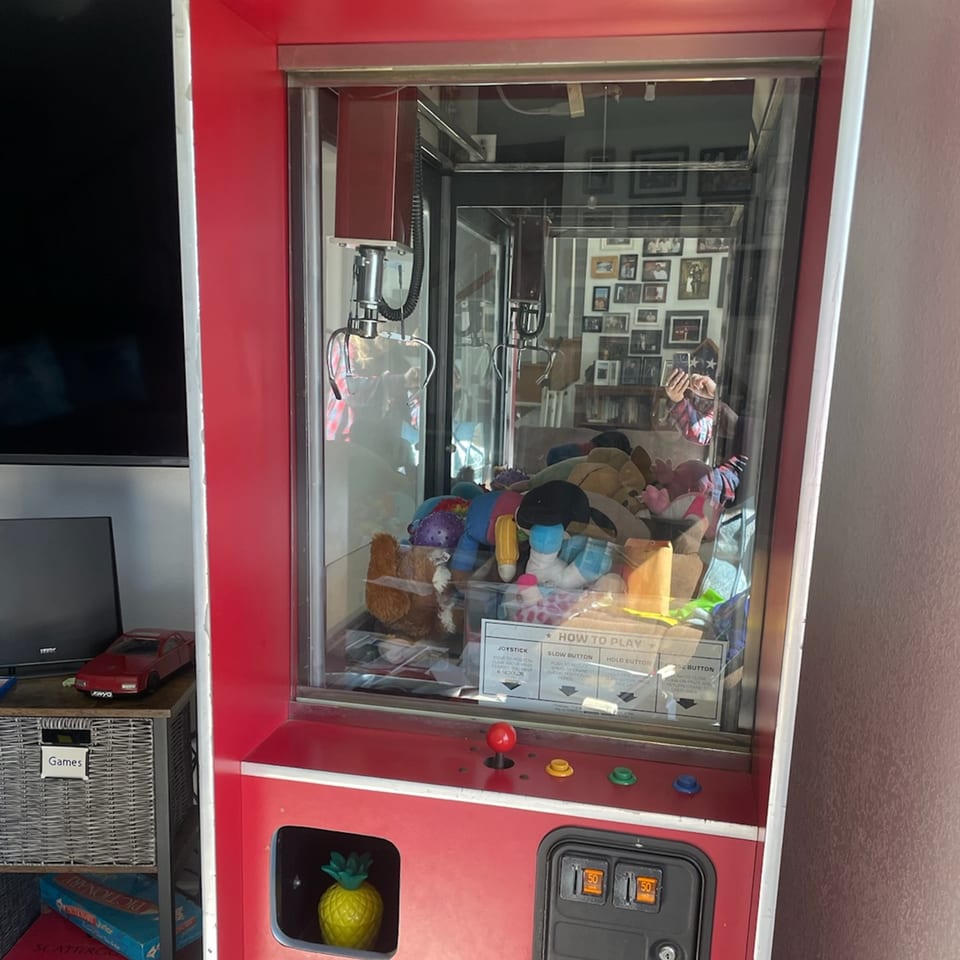 A red claw machine filled with stuffed animals, next to a shelf with board games.