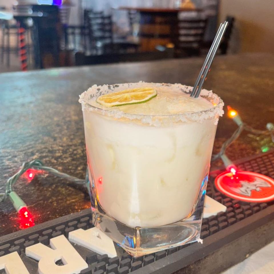 A frosty margarita with a lime slice and metal straw sits on a bar with string lights.